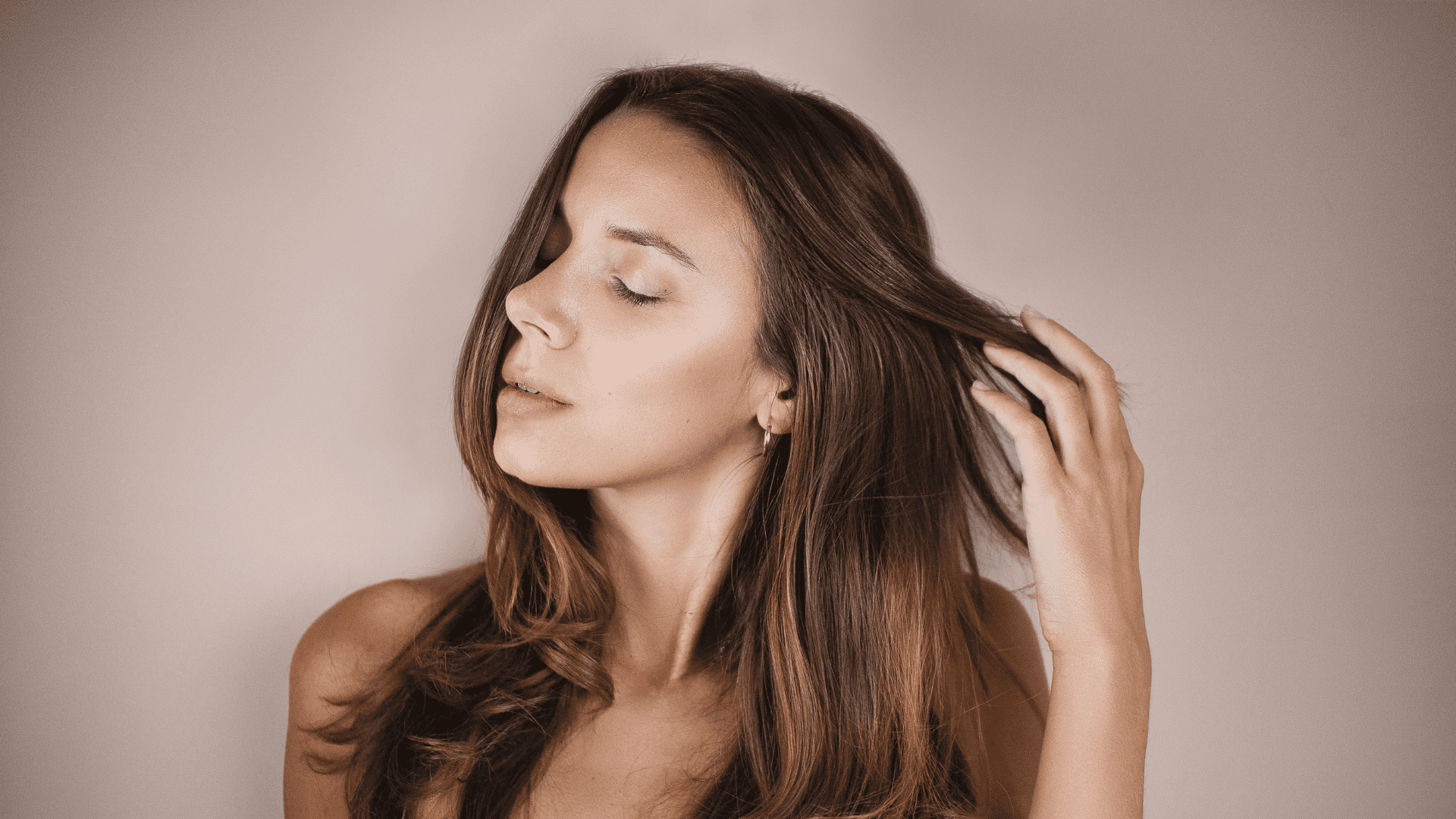 Woman with brown hair touching her hair and looking serene against a neutral background.