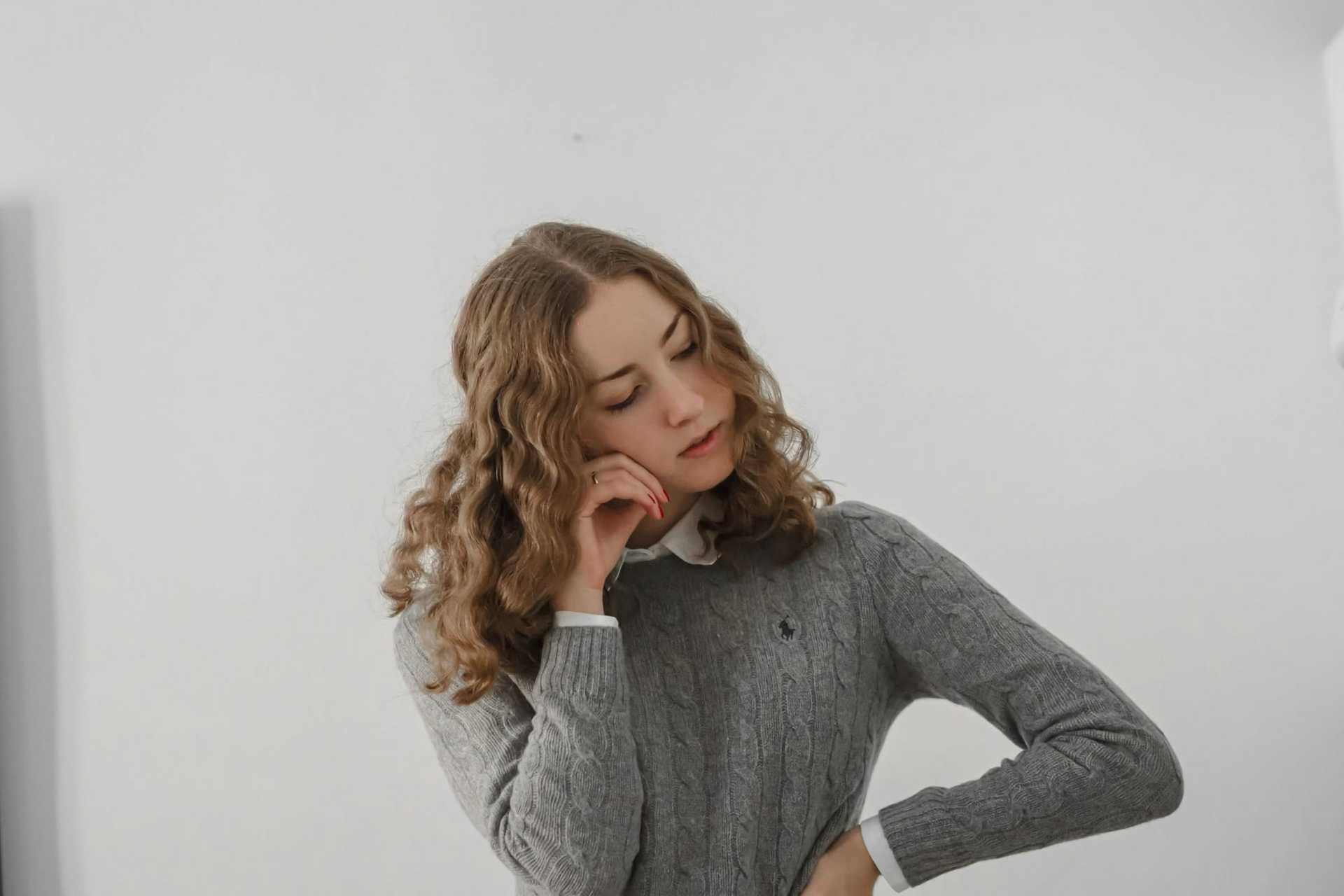 Young woman in a gray sweater, with curly hair, resting her head on her hand thoughtfully.